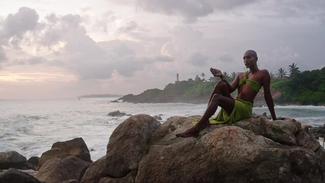 Divine non-binary black person in long open dress brass jewelry poses on rocky hill top above dramatic ocean sunset skyline. Queer lgbtq fashion model in open outfit sits on hill. Pantheon concept