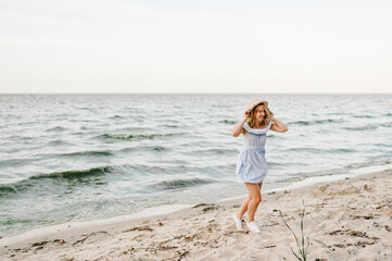 Happy beautiful girl running on beach ocean and enjoying sunny summer day on vacation. Portrait of stylish female on sand. Young attractive smiling blond woman in a straw hat joyfully walks near sea.