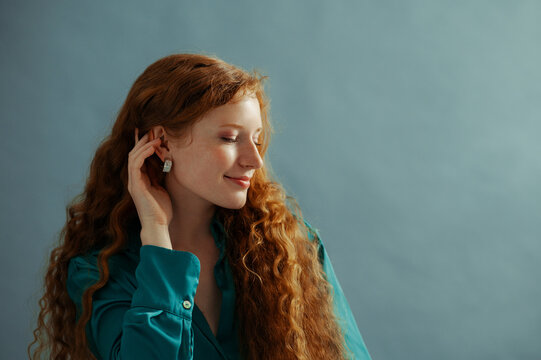 Beautiful happy smiling redhead freckled woman with long curly hair, wearing trendy silver earrings, posing on blue background. Close up studio portrait. Copy, empty, blank space for text

