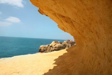 A rocky cliff with a sandy beach leading to it. The ocean is blue and calm. The sky is blue with a few clouds.