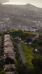 A vertical top-down shot captures houses perched on a hill overlooking Funchal city and the Atlantic Ocean in the background. Enhanced by the presence of a white car driving along the road.