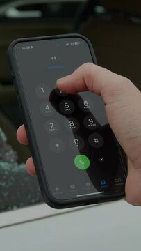 Hand dials 112 on smartphone after spotting shattered car window instant reaction to emergency vertical video. Fingers tap 112 mobile screen upon noticing broken vehicle glass quick response crisis.