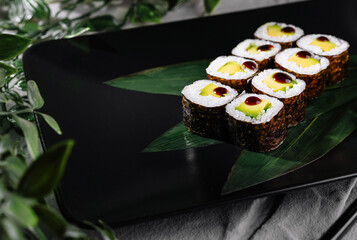 Sushi rolls garnished with wasabi and ginger on a modern black plate, botanical backdrop