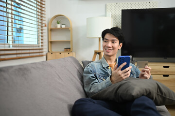 Smiling young man with mobile phone and credit card looking away while sitting on sofa