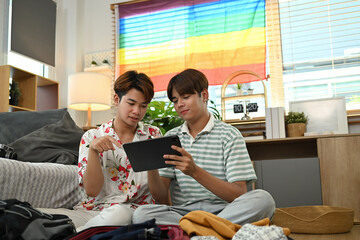 Young gay couple booking hotel reservation on digital tablet and packing luggage to travel vacation