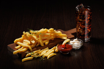 A pile of French fries served on a wooden board with mayonnaise and ketchup. Cola with ice in a...