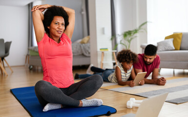 Young woman, mother exercising at home in living room, father playing with kid in background.