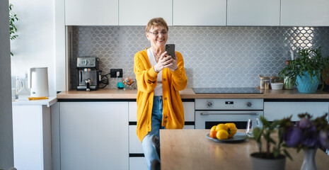 Elderly attractive happy woman standing in the kitchen with a smartphone in her hands, online at home
