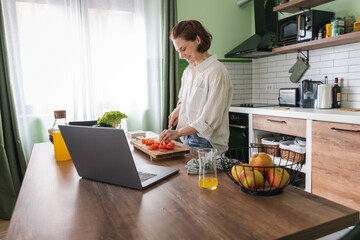 Young happy woman standing in the kitchen preparing salad and communicating online using laptop