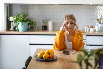 Senior Caucasian woman sitting at the table in the kitchen holding her head, headaches concept