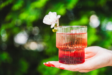 A hand holds a glass of pink beverage with a white flower garnish against a blurred green background