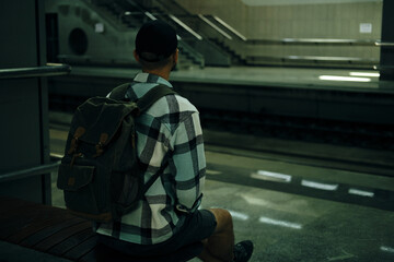 A young man travels alone with a backpack. The guy is sitting on a bench underground and waiting for his train. Rear view. A railway station in Belgrade, Serbia.