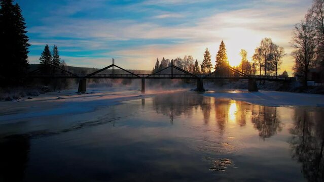 Drone of idyllic view of a bridge on frozen Glomma River with trees in the background at sunset