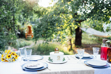 Cute baby birthday cake with number two and lion cub on holiday table outdoors. Nobody here. Laid festive table with flowers, tablecloth, plates and glasses in cozy place in backyard of house.
