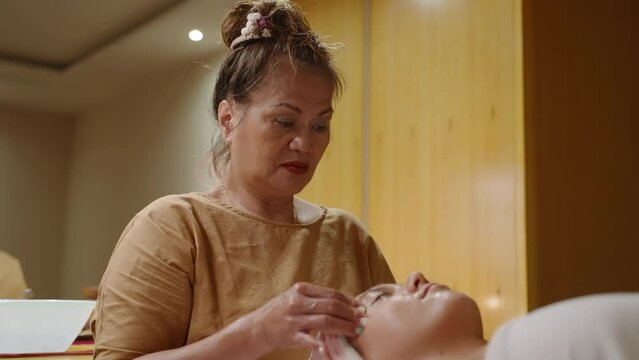 Senior asian masseuse massaging a young female client face with hands using oil in spa. Adult woman worker of a wellness center gives relaxing facial massage to a tanned girl, presses trigger points
