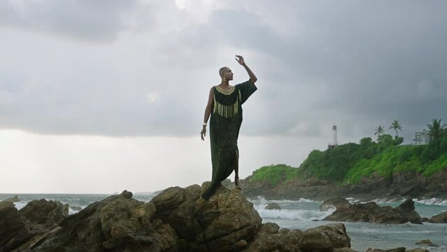 Gay bipoc man in luxury dress, boutique brass jewelry, makeup poses gracefully tropical rocky hill top on ocean beach. Homosexual ethnic elegant person standing on stone like Oshun orisha. Cloudy sky.