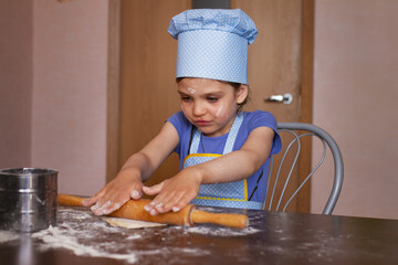 Portrait of 5 year old girl in blue dress and dressed as chef in kitchen laughs and rolling out...