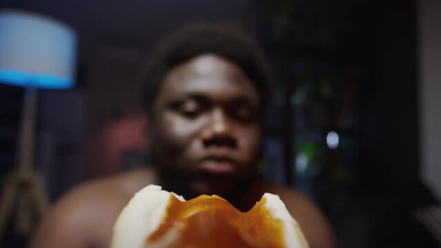 Close up POV shot of hungry Black man with overweight eating hot dog during night at home