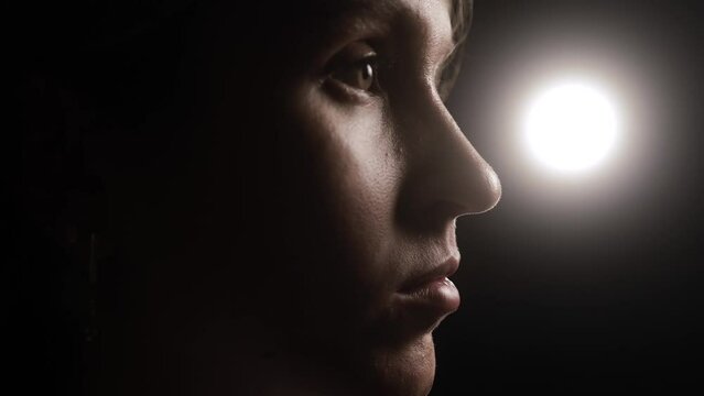 Thoughtful face of woman against dark background in spotlight. Camera moves around face and bright bokeh appears with beautiful halo bokeh
