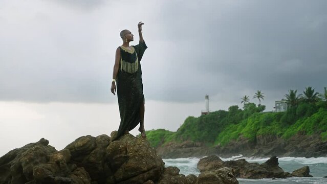 Divine non-binary black person in boutique dress brass jewelry poses on rocky hill top above dramatic ocean cloudy skyline. Queer lgbtq fashion model in posh outfit fluttering in air. Pantheon concept
