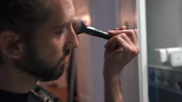Young gay man puts on makeup looking in the mirror at home
