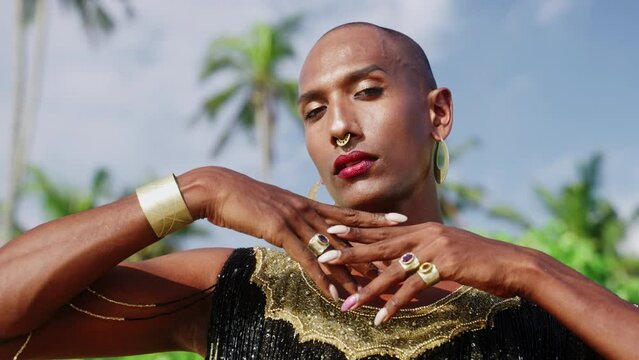 Gay bipoc man in luxury dress boutique brass jewelry, makeup poses looking on camera with moody face. Homosexual ethnic elegant person in posh gown wears nose-ring piercing, earrings. Palm trees.