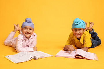 Two Happy indian sikh school boys lying on floor with note books studying isolated over yellow...