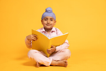 Portrait of smiling indian sikh school kid sitting on floor holding note book studying isolated...