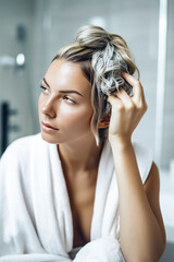 Fototapeta premium A woman with blonde hair is sitting in a bathtub with a towel draped over her shoulders. She is looking at her reflection in the mirror