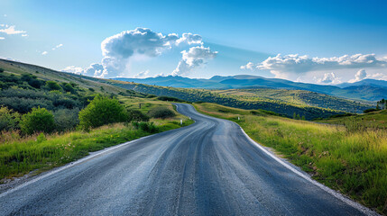 Naklejka premium Winding road through green hills. Scenic view of a winding asphalt road through lush green hills, leading towards distant mountains under a bright blue sky with fluffy white clouds.