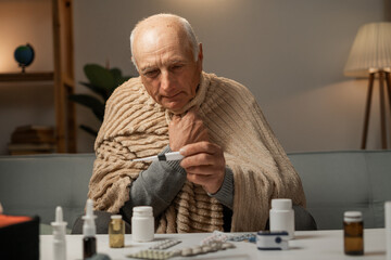 elderly man, wrapped in blanket, measures his temperature. Self-isolation and sick leave home. medicines