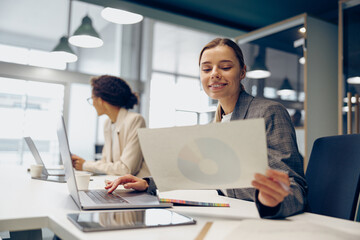Smiling female entrepreneur working with documents and use laptop while sitting the desk in office 