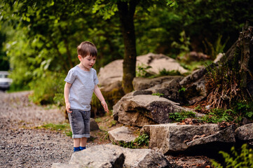 Young boy in a gray t-shirt and denim shorts exploring a gravel path near large rocks and trees. Capturing the essence of childhood curiosity and adventure in nature.