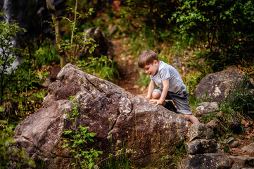 Smiling young boy in a gray t-shirt and denim shorts climbing large rocks in a forested area. Capturing a moment of joyful adventure and exploration in nature.