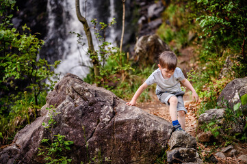 Smiling young boy in a gray t-shirt and denim shorts climbing large rocks in a forested area....