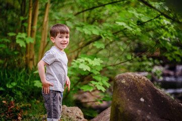 A young boy smiles brightly while standing on a forest path surrounded by lush green foliage and rocks. He is enjoying a peaceful nature walk.