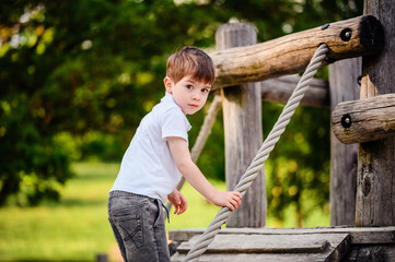 A young boy in a white shirt and jeans climbs a wooden playground structure using a rope, set against a backdrop of green trees and grass on a sunny day.