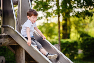 A happy young boy slides down a metal playground slide on a sunny day, surrounded by lush green trees, enjoying outdoor playtime. © Oleksii Yemelianov