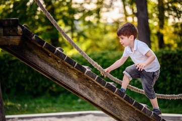 A young boy in a white shirt and denim shorts carefully climbs a wooden ramp using a rope at a...
