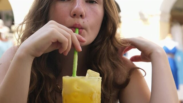 A girl happily spits freshly squeezed orange juice through a straw. European woman relaxes and travels to warm countries.