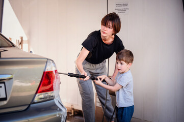 Mother closely assisting her young son as they wash the rear of a car at a self-service station....