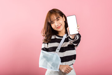 Naklejka premium Happy Asian woman with a broken arm, wearing an arm splint, smiles while displaying her smartphone screen. Coping with pain and recovery, isolated on pink.