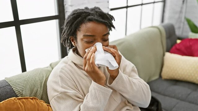 A young african american woman with dreadlocks blows her nose on a sofa indoors, suggesting illness or allergies.