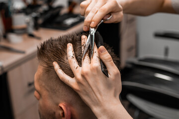 Professional barber styling hair of his client in the barbershop using scissors and comb.