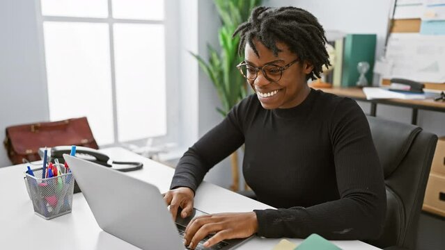 Cool and confident, a young black woman proudly sporting dreadlocks, working, smiling with gleaming teeth at laptop in office - epitome of joyful success!