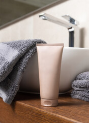 Beige cosmetic tube near grey folded towels on basin on wooden countertop in bath, mockup