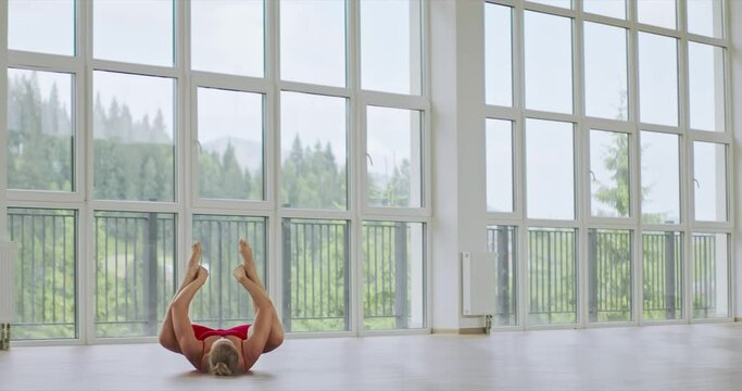 Flexible woman exercising, stretching legs, while lying on floor in studio. Back view of graceful female wearing red activewear doing in leg-split, against panoramic window, in motion. Yoga concept.