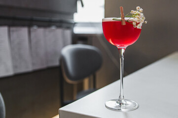  vibrant red cocktail in a stemmed glass, adorned with a sprig and flowers, rests on a bar counter with a soft-focus background
