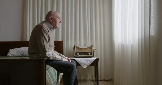 A senior man sits on his bed, beside a large window, gazing thoughtfully at a vintage radio in a peaceful room.