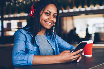 Portrait of attractive dark skinned hipster girl listening to music in headphones using application with favorite playlist.Attractive afro american student resting in coffee shop texting messages